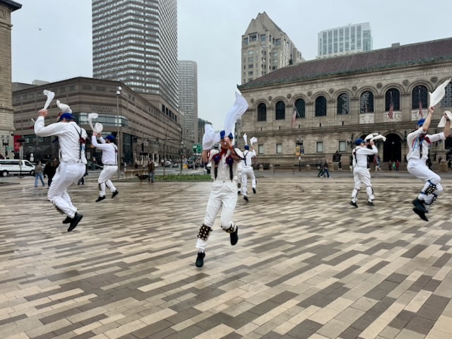 Newtowne Morris performing Ducklington The Fox on Copley Plaza across from the Boston Public Library, the dancers in whites, suspenders, and blue caps forming a circle and leaping into capers facing out.