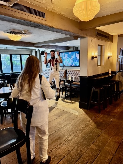 A Morris dancer in whites and suspenders dances in an empty bar with a TV behind on the right and windows on the left, the musician in whites stands in the foreground with her back to the camera.