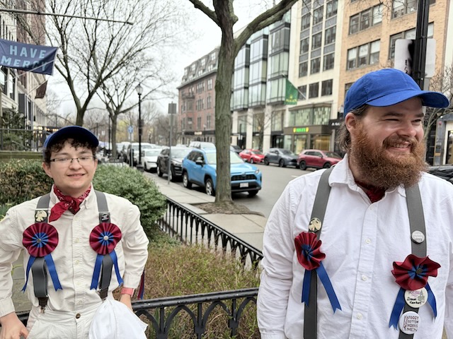 View of Newbury Street with two people in Morris kits, whites, gray suspenders with rosettes, and blue caps, standing in front of a metal railing.