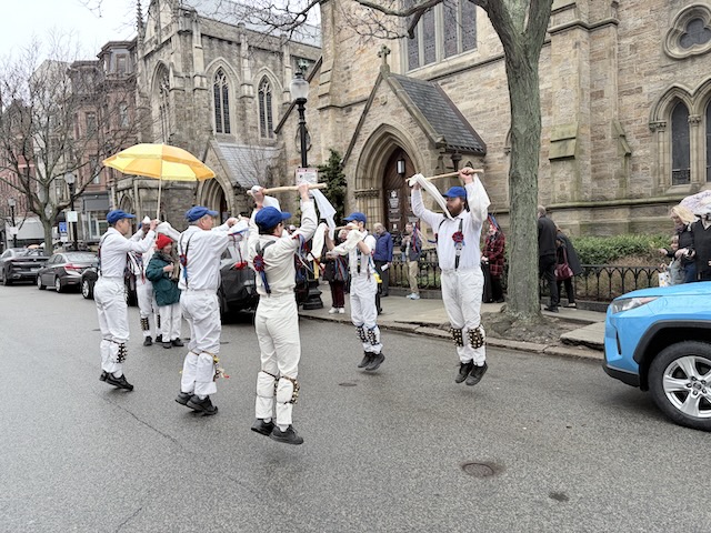 Newtowne Morris performing Stanton Harcourt Nightingale on Newbury Street in front of Emmanuel Church, two lines of dancers in whites, suspenders, and blue caps holding sticks abover their heads and jumping into the air.