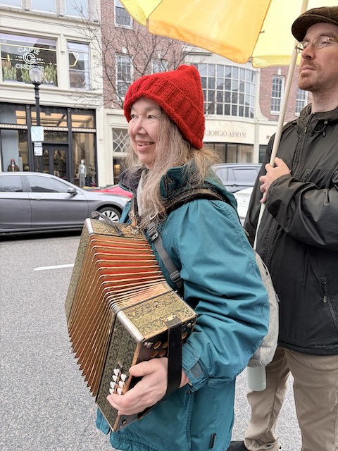 Musician wearing a red knit cap and holding a melodeon standing on a city street under a yellow umbrella being held by a person standing behind.