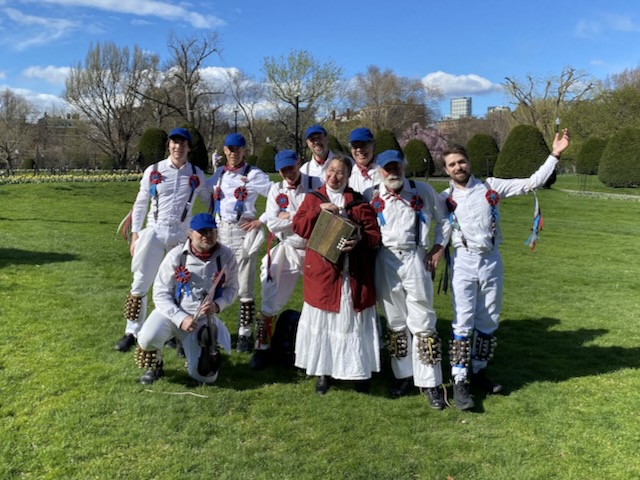 Nine members of Newtowne Morris standing on a lawn in the Public Garden wearing whites, bells, suspenders, and blue caps, with the musiciain in the center in a red coat holding a melodeon.